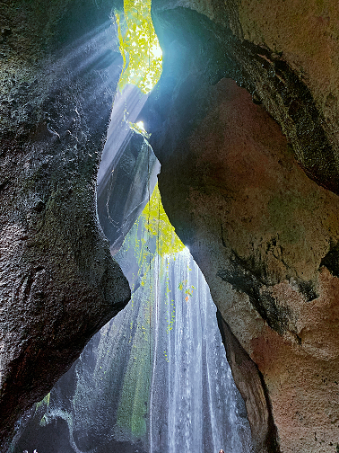 A captivating view from inside a cave looking up at a narrow opening where sunlight streams in, illuminating the cascading waterfall and the vibrant green foliage that peeks through, creating a magical and almost otherworldly atmosphere