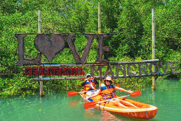 The image depicts two individuals kayaking in a calm mangrove waterway. They are both wearing bright orange life vests, with the person in front raising a paddle in the air in a cheerful manner. The kayak is orange, complementing the vibrant green of the mangrove trees surrounding the water. Behind them is a large, rustic sign featuring the word "LOVE" with a heart symbol replacing the 'O', followed by the word "MANGROVE". The sign appears weathered and is mounted on tall wooden posts, suggesting a natural, outdoor adventure setting. The sky is clear, hinting at a sunny day, perfect for water activities.