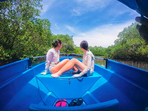 Two visitors seated in a vibrant blue glass-bottom boat, gazing into the clear waters of Bali's mangrove forest, surrounded by verdant mangrove trees.