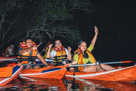 Close-up of smiling kayakers in life jackets, enjoying a night tour in Bali's mangrove forest, with one person waving cheerfully.