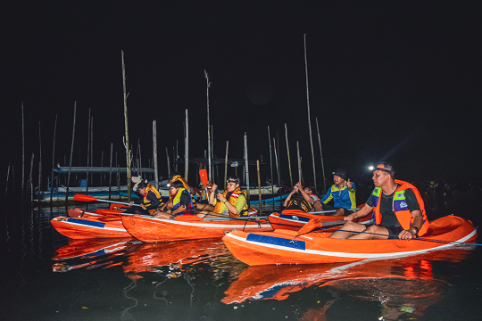 Kayakers captured from a distance, paddling through the dark waters of Bali's mangrove forest with headlamps and boat lights reflecting on the water.