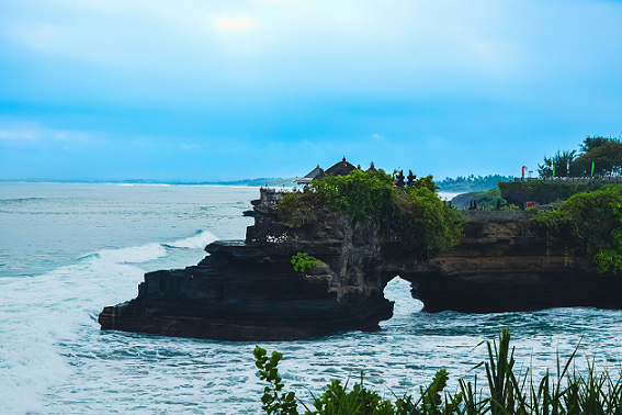 A rocky cliff jutting into the sea with a traditional Balinese temple perched on the edge, under a cloudy sky, surrounded by turbulent waves.