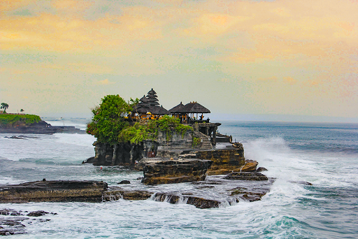 The temple on a rocky cliff with waves breaking against it, a clear view of the temple's multi-tiered thatched roofs, and trees, under a cloudy, twilight sky.