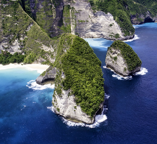 An aerial view of Kelingking Beach in Bali, Indonesia, showcasing the iconic T-Rex-shaped cliff covered in lush greenery, flanked by pristine white sandy beaches and clear turquoise waters, with waves gently lapping against the shore.