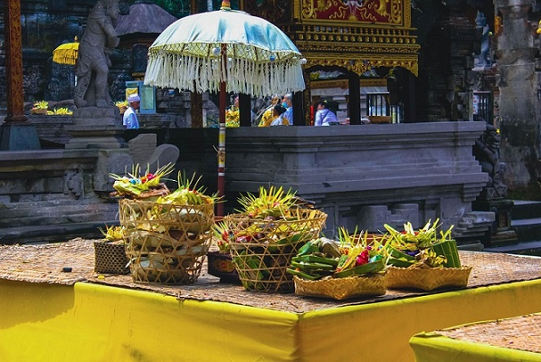 This image shows traditional offerings, which are placed on a yellow cloth. The offerings are in woven baskets and decorated with a variety of items, including flowers. In the background, there’s a structure with Balinese architecture featuring intricate details and a parasol, suggesting this is a temple or a ceremonial place. It seems to be a sunny day, and there are a few people in the distance, which adds to the peaceful and spiritual ambiance of the scene. This setting is typical for Bali, Indonesia, where such offerings are a part of daily religious practices.