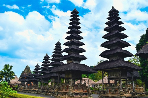 The image features the iconic meru towers at Taman Ayun Temple, each tiered roof reaching high into the sky. These multi-layered towers, made from wood and thatched roofs, symbolize sacred mountains in Hinduism and are dedicated to various deities. The intricate stone carvings at the base of the towers complement the spiritual significance of the site, while the surrounding greenery adds to the temple’s peaceful atmosphere. The clear blue sky in the background enhances the majestic presence of the meru towers, making this a beautiful and serene scene.