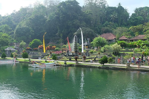 A serene scene at Tirta Gangga Water Palace featuring a large pond with small boats docked by the shore. The 11-tiered fountain stands tall in the background, surrounded by vibrant gardens and colorful flags, with visitors exploring the lush landscape and peaceful surroundings.