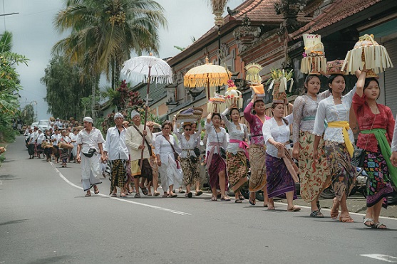 A group of Balinese women in traditional attire walk in a ceremonial procession, carrying offerings on their heads as part of a religious event. The street is lined with spectators, creating a festive atmosphere