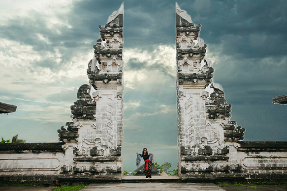 A person stands at the iconic split gate of Lempuyang Temple, dressed in traditional Balinese attire, against a dramatic sky filled with clouds. The temple’s intricate architecture frames the figure, highlighting the cultural significance of this sacred site.