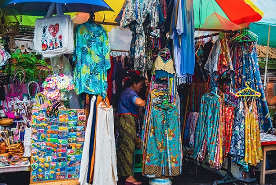 A vibrant market stall in Bali displaying a variety of colorful clothing, bags, and souvenirs under a rainbow-colored umbrella, with a local vendor arranging items.