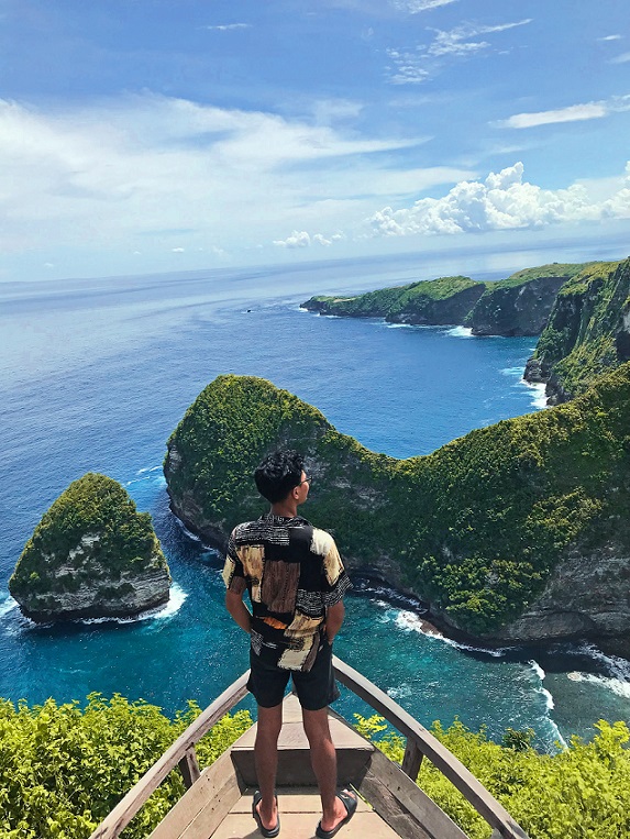 A visitor stands on a wooden lookout platform, taking in the breathtaking view of the rugged cliffs, turquoise waters, and lush green landscape of Kelingking Beach on Nusa Penida Island, Bali.