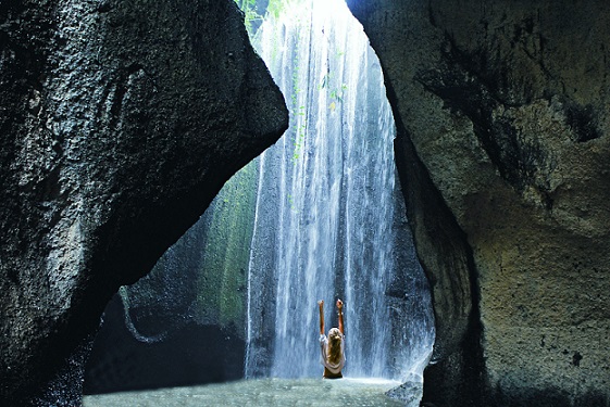 Traveler standing in awe beneath a cascading waterfall, surrounded by towering rock formations and a serene, hidden grotto in Bali