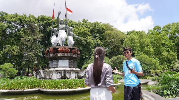 A tour guide is enthusiastically explaining the history of the Puputan Badung Monument to a female tourist in Bali. The statue, draped in white cloth with Indonesian flags, stands tall surrounded by lush greenery and a peaceful water feature.